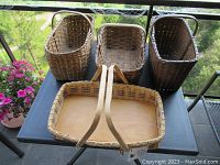 Four assorted baskets shown on black table outdoors with greenery in background. Includes 3 antique woven baskets and 1 flat wooden basket with handles.