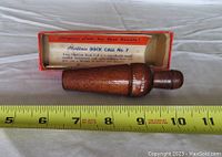 Photo of the wooden duck call next to a measuring tape showing approximately 5 inches in length, with the original box in the background.