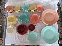 Top-down photo of all cups, saucers, and plates in multiple colors (coral, aqua, yellow, burgundy, white) arranged on white cloth