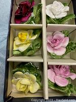 Top view of six small floral candle holders with yellow, pink, white, and burgundy roses over green leaves, displayed in a cardboard box.