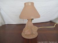 Photo showing pink glass lamp with seated figure base and textured pink lampshade on wooden surface against white backdrop.