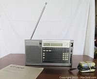 Front view of Sony ICF-2010 radio with antenna extended, showing speaker, digital display, buttons, manual, and adapter on table