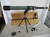 Mirador telescope fully assembled on a small black tripod with additional eyepieces and lens cap on table beside it. Wooden storage box open behind the telescope.