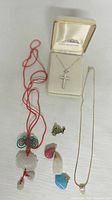 Photo showing three necklaces including sterling silver cross necklace in box, red cord necklace with carved pendant, gold-tone chain necklace with small star pendant and four small pendants with colorful and metal finishes