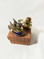 Two cloisonné enamel brass duck-shaped lidded trinket boxes displayed on a patterned box, showing detailed enamel and brass work.