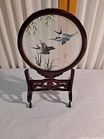 Front view of round shadow box on carved wooden stand showing birds flying over pond scene