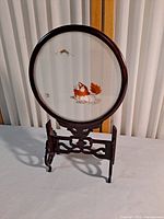 Front view of round shadow box showing detailed fabric artwork with dragon, orange leaves, and mountain scene inside dark wood circular frame with carved wood stand.