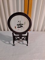 Front view of round shadow box with panda painting and wood stand on white surface against wooden backdrop.