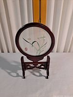 Front view of round shadow box with yellow insect and white flowers inside wooden frame on carved stand.