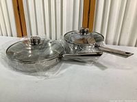 10-inch skillet and 8-inch saucepan both with clear glass lids and metal handles, wrapped in plastic packaging.