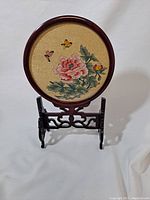 Front view of round shadow box with embroidered pink flower and butterflies, displayed on carved wooden stand.