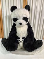 Large black and white stuffed panda holding a smaller panda on lap, sitting position against vertical backdrop.