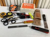 Photo showing tape measures, multiple scissors including Spear brand trip scissors in box, multi-bit screwdriver set, a yellow-handled screwdriver, and a metal file arranged on a white surface.