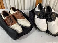 Two pairs of FootJoy golf shoes placed on storage bags, one pair white and brown woven texture, the other white and black with scuffs and signs of wear.