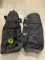 Two black golf bag carriers seen top-down on a concrete floor, showing wheels, straps, and zippered pockets.