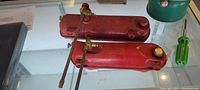 Two red Coleman stove gas tanks with brass valves and control rods, showing tops and valves, against white background.