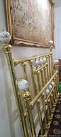 Photo showing full view of the brass bed frames leaning against wall under a large framed tapestry. Clear view of the brass frame and porcelain floral finials.