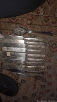 Full set of antique silver-plated silverware laid out on a carpet background. Includes knives, serving spoon, ladle, and fork.