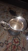 Silver plated creamer pitcher showing ornate handle and tarnished surface on a patterned carpet.