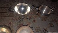 Top view of four silver plated pieces on a patterned carpet: a handled bowl, creamer, sugar bowl with lid, coffee pot.