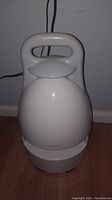 Photo of white portable speaker, front angle showing top handle, plugged in, on floor near wall.