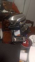 Image showing the full waffle iron with the lid open, held by a hand, with a table and chairs in background.