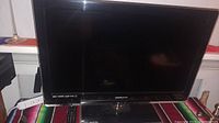 Image of the front view of a Hannspree flat-screen television on a black base, placed on a colorful striped cloth with a remote visible underneath the screen.