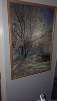 Framed picture showing a snowy winter landscape with trees in a light wooden frame, viewed hanging on a wall.