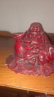 Front and side view of red Buddha figurine illustrating facial details, seated posture, and the coins around the base.