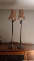 Pair of small vintage table lamps with bronze/dark metal bases and beige fabric shades with beaded fringe, placed on wooden furniture.