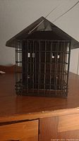 Front view of black metal bird feeder cage shaped like a small house with wire hanging loop.