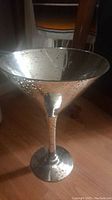 Front angled view showing the large metallic martini glass with speckled distressed finish, wide bowl shape, and long stem on wooden floor with some furniture background.
