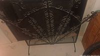 Front view of wrought iron decorative fan-shaped fireplace screen showing intricate scroll and leaf design and mesh backdrop.