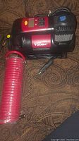 Front view of the black and red portable air compressor with attached coiled red hose and power cord with cigarette lighter plug, placed on a patterned surface.