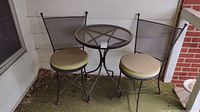 Full set including the table and two chairs with beige cushions placed on porch area, close to a wall.