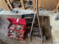Two red metal toolboxes with assorted hand tools on garage floor alongside two large wooden-handled rusted hand tools