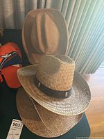 Stacked cowboy hats including straw hats with decorative leather bands and several felt and leatherlike hats in brown and beige tones