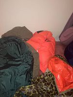 Pile of hunting jackets including hunter orange and camo with orange lining, wool pants visible beneath.
