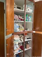 Wooden linen closet with multiple shelves containing folded towels, linens, cleaning sprays, and bathroom supplies.