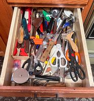 Top view of an open kitchen drawer containing assorted kitchen utensils including scissors, a pizza cutter, wooden handled spoons, ladles, can openers, and miscellaneous small utensils in plastic and metal.