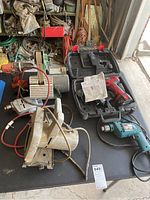 Overview of assorted power tools on black table including belt sander, circular saw, blue corded drill, and Skil cordless drill in case.