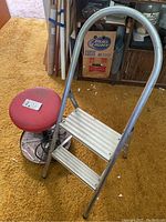 Photo of aluminum two-step ladder alongside a red cushioned rolling garage seat on a brown carpet.