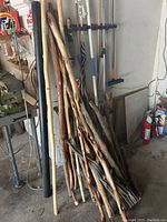 Five walking sticks with hand-carved designs, rough textures, leaning against a wall along with other items in garage setting.