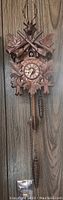 Full view of wooden cuckoo clock with carved leaves, deer head, crossed rifles, pinecone weights, and clock face with roman numerals