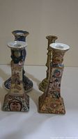 View of four candle holders, three porcelain with intricate Asian hand-painted designs, one brass holder in background.