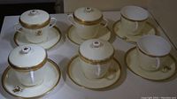 6 porcelain teacups and 6 matching saucers displayed on table. 4 cups have lids, 2 cups do not. White with gold trim and floral emblem.