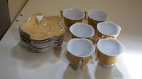Photo showing six white ceramic teacups with gold floral patterns and handles alongside six matching saucers stacked.