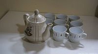 Photo showing full set of ceramic teapot and eight cups arranged behind it on a white surface.