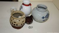 Six porcelain items arranged on a white surface: a beige candle holder with holes, a small dark brown cup, a gray vase with floral patterns, a white sake bottle with red string, two small white cups with Japanese writing, and a tiny gray figurine.
