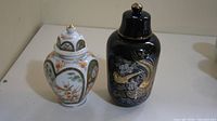 Pair of two lidded vases on a light surface showing contrasting designs — one white with floral patterns and one black with a dragon motif.